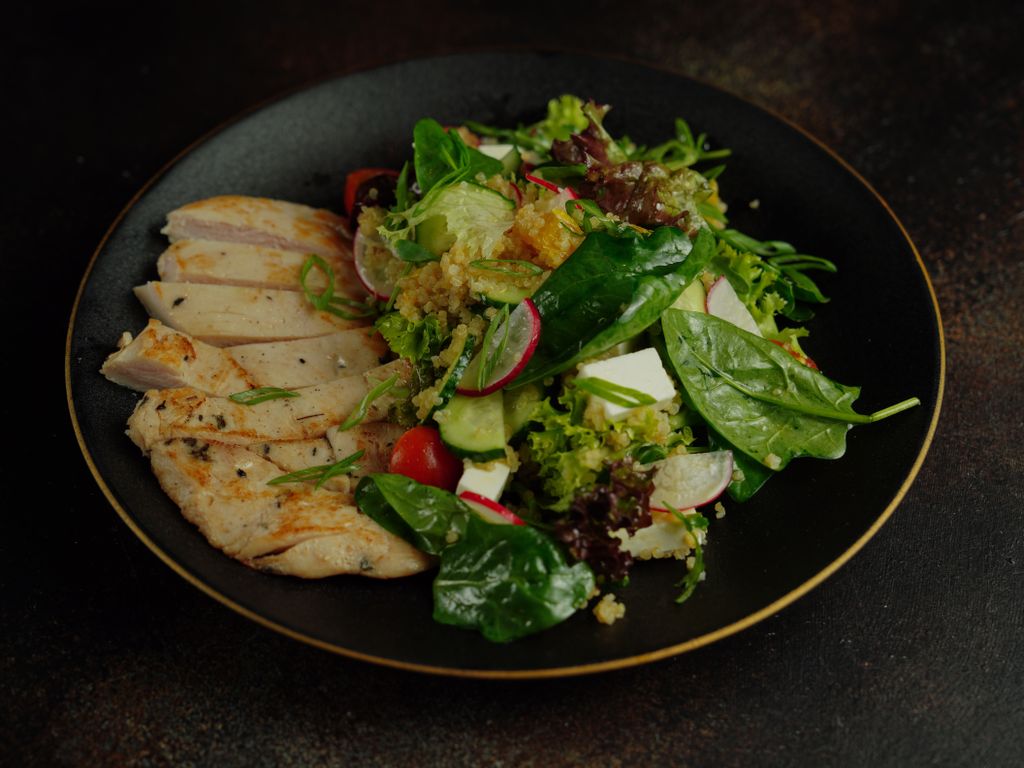 Photo by Nadin Sh A vibrant salad featuring grilled chicken, quinoa, and fresh vegetables on a black plate.
