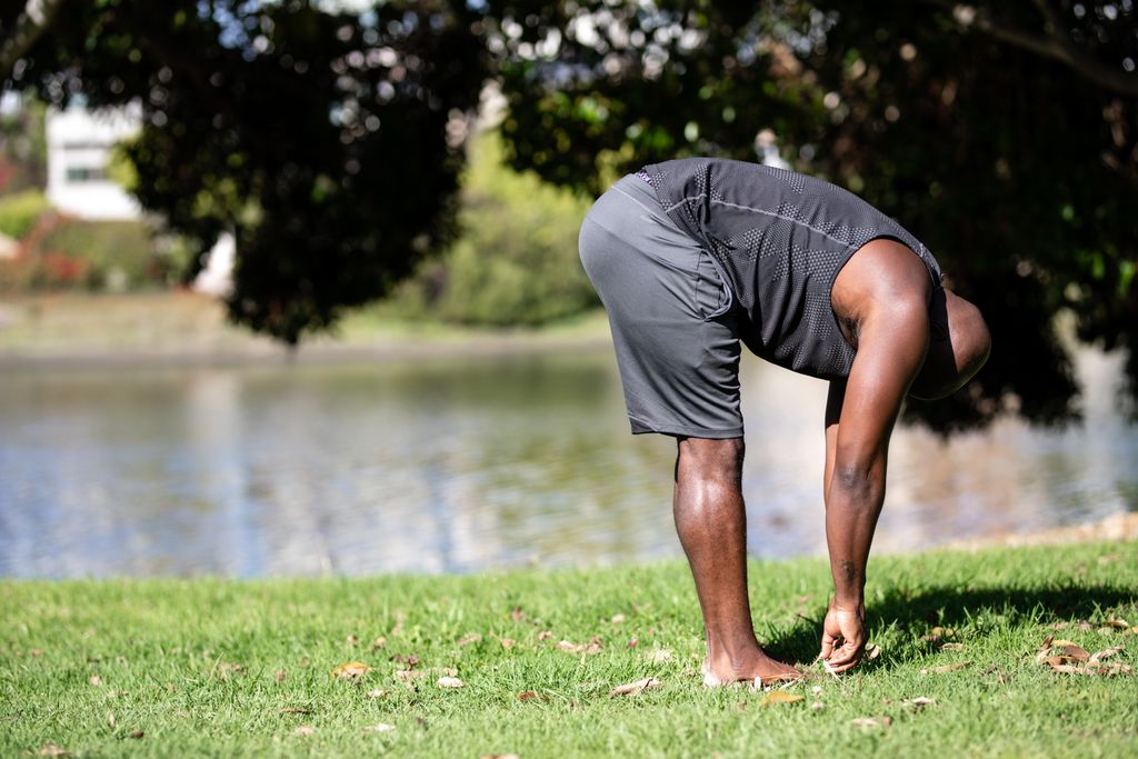 Athletic man in a park stretching near a lake during summer. Promotes fitness and relaxation.