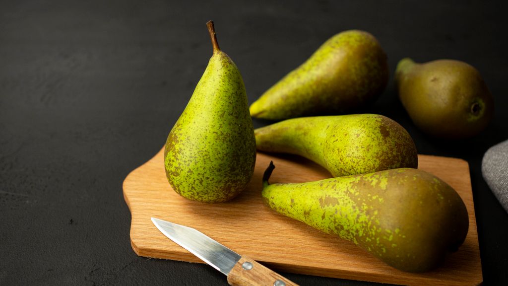 Photo by Mateusz Feliksik Four ripe green pears on a wooden board with a knife on a dark background.